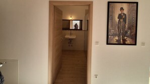Une pièce avec un plafond haut, des poutres en bois, un sol carrelé, une salle de bain avec un lavabo et un tableau encadré au mur.