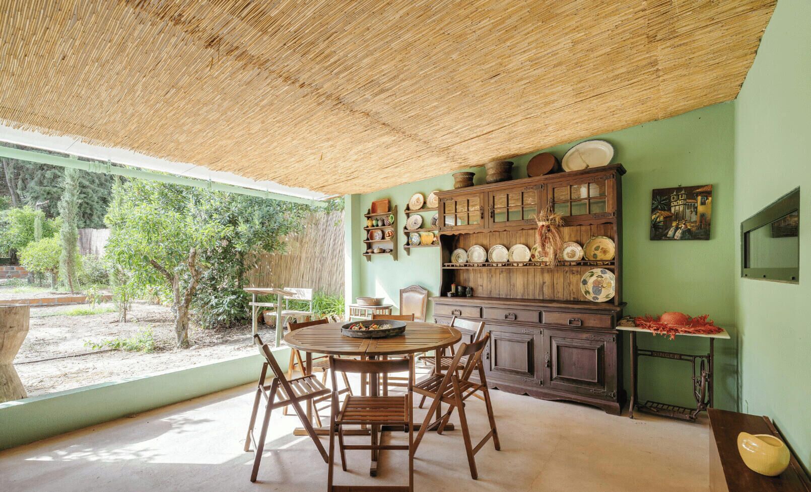 A dining area with a round wooden table and chairs, a kitchen with wooden cabinets and a hanging pot rack, and a view of greenery outside.