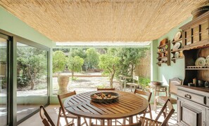 Un espace repas comprenant une table ronde en bois, des chaises, un plafond en osier et une vue sur des arbres et un jardin.