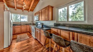 A kitchen with wooden cabinets, a granite countertop, and a large window with a view of trees. - Lake Tahoe Tree House with Panoramic Views of Lake Tahoe (Zephyr Cove)