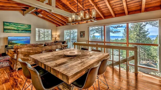 A spacious room with a large wooden dining table, comfortable seating, and large windows offering a scenic view. - Lake Tahoe Tree House with Panoramic Views of Lake Tahoe (Zephyr Cove)