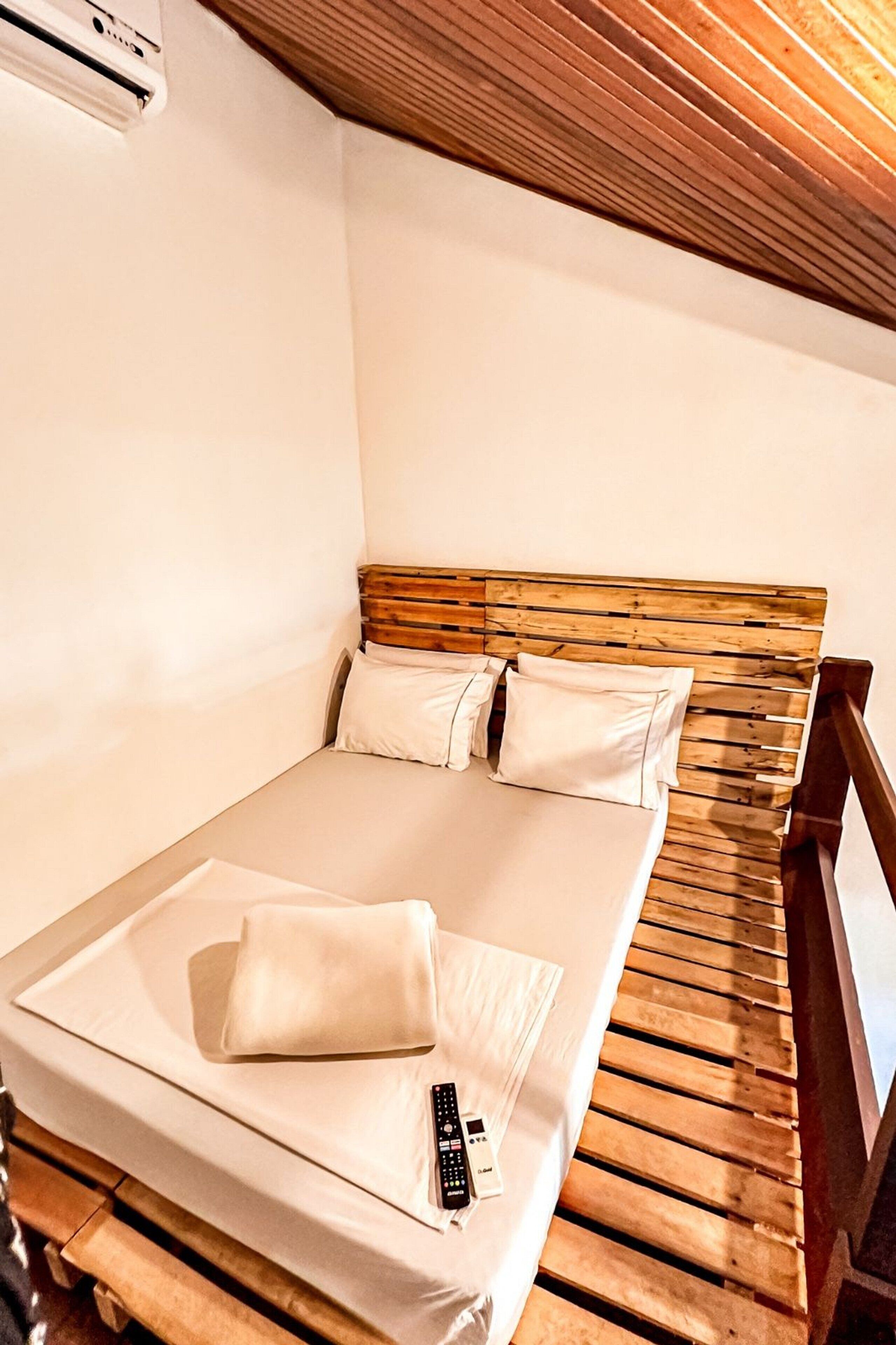 Un lit simple avec une tête de lit en bois, une literie blanche et une télécommande posée sur le lit.