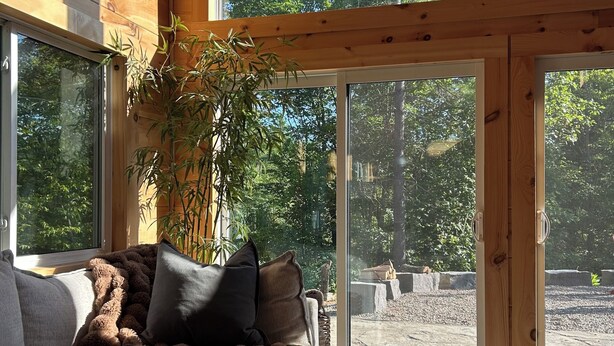 Tunnelmallinen huone, jossa on puinen sisustus, suuret ikkunat, peitolla varustettu sohva ja ruukkukasvi.