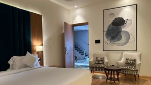 Une chambre d’hôtel avec un grand lit, deux fauteuils, une table ronde et une peinture abstraite moderne accrochée au mur.