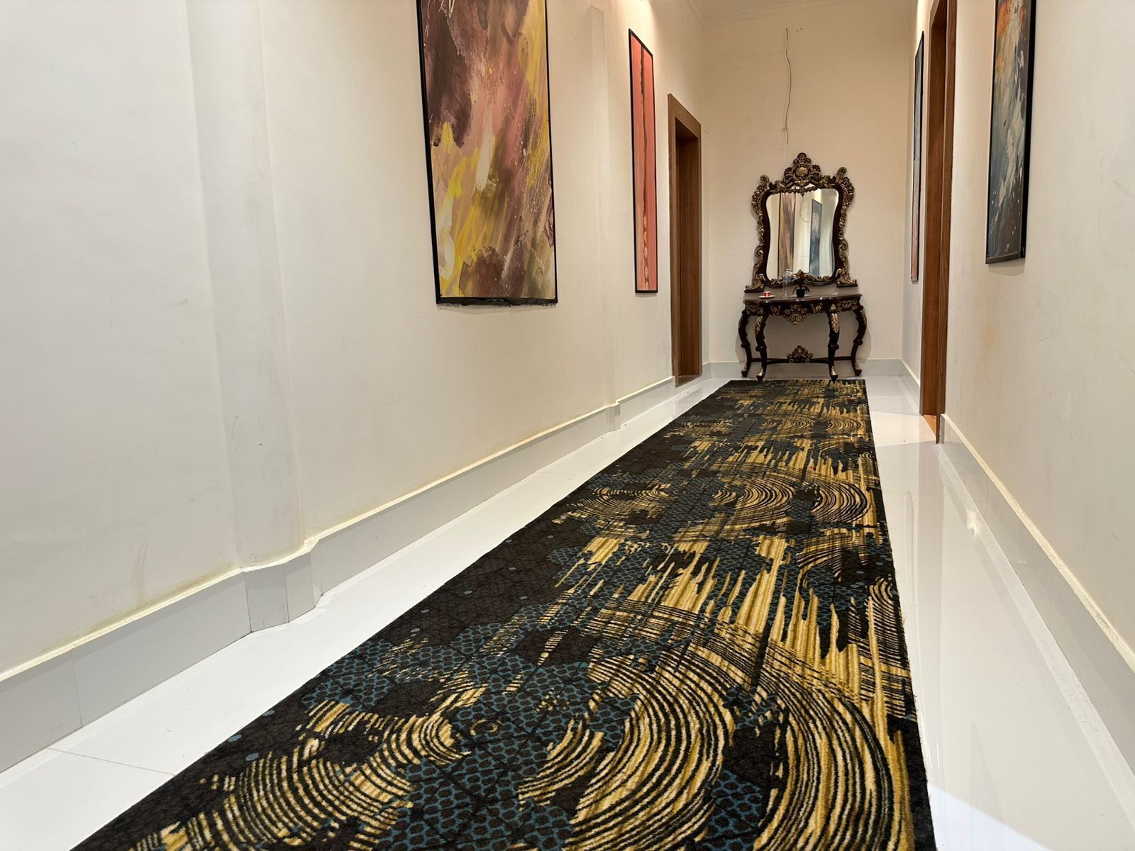 Un pasillo con alfombra estampada, obras de arte enmarcadas en las paredes y un espejo sobre una mesa.