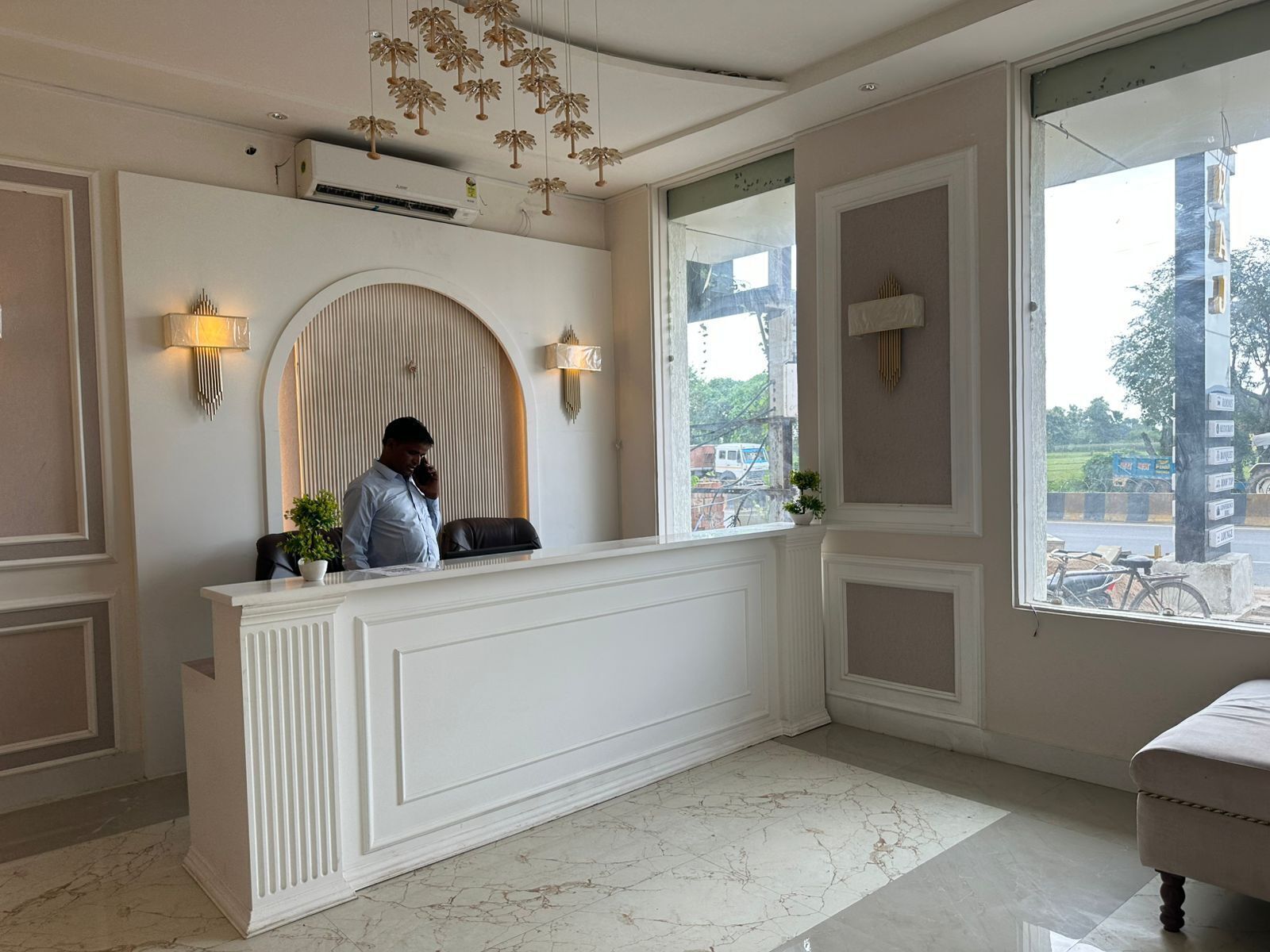 A reception desk with a man standing behind it, a large arched mirror, and a view of the street outside.