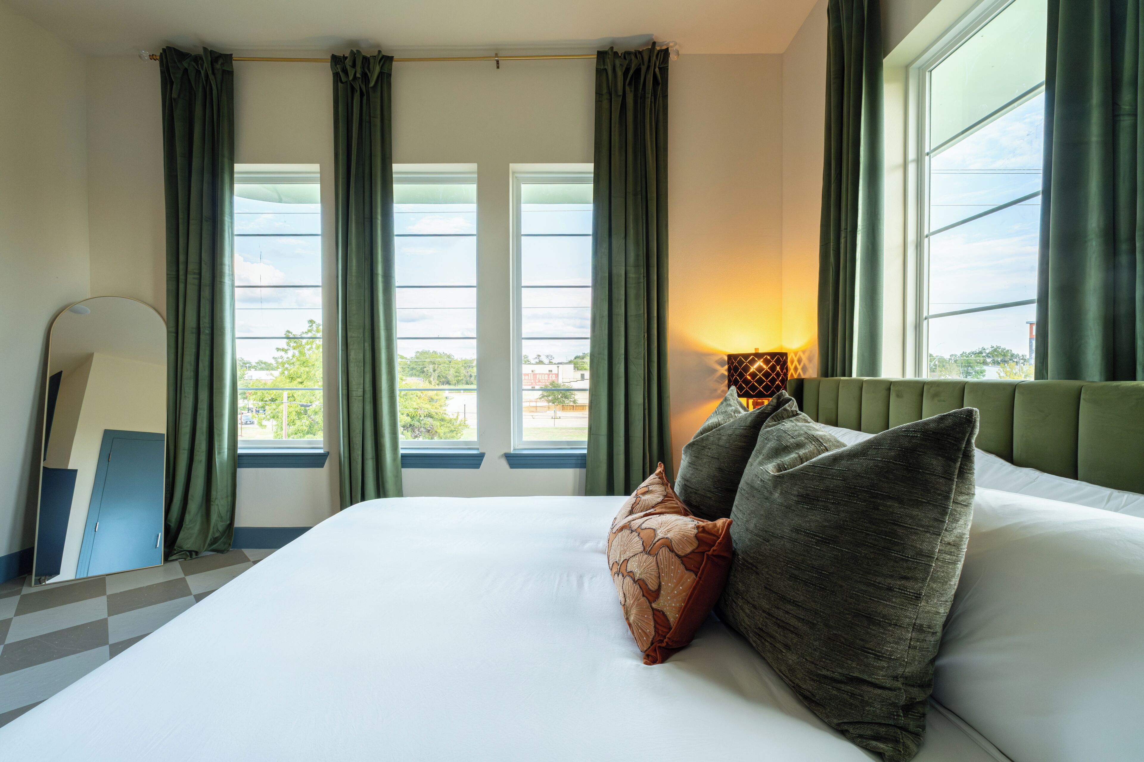 Habitación de hotel con una cama grande, dos ventanas con cortinas, una lámpara de noche y vistas al exterior.