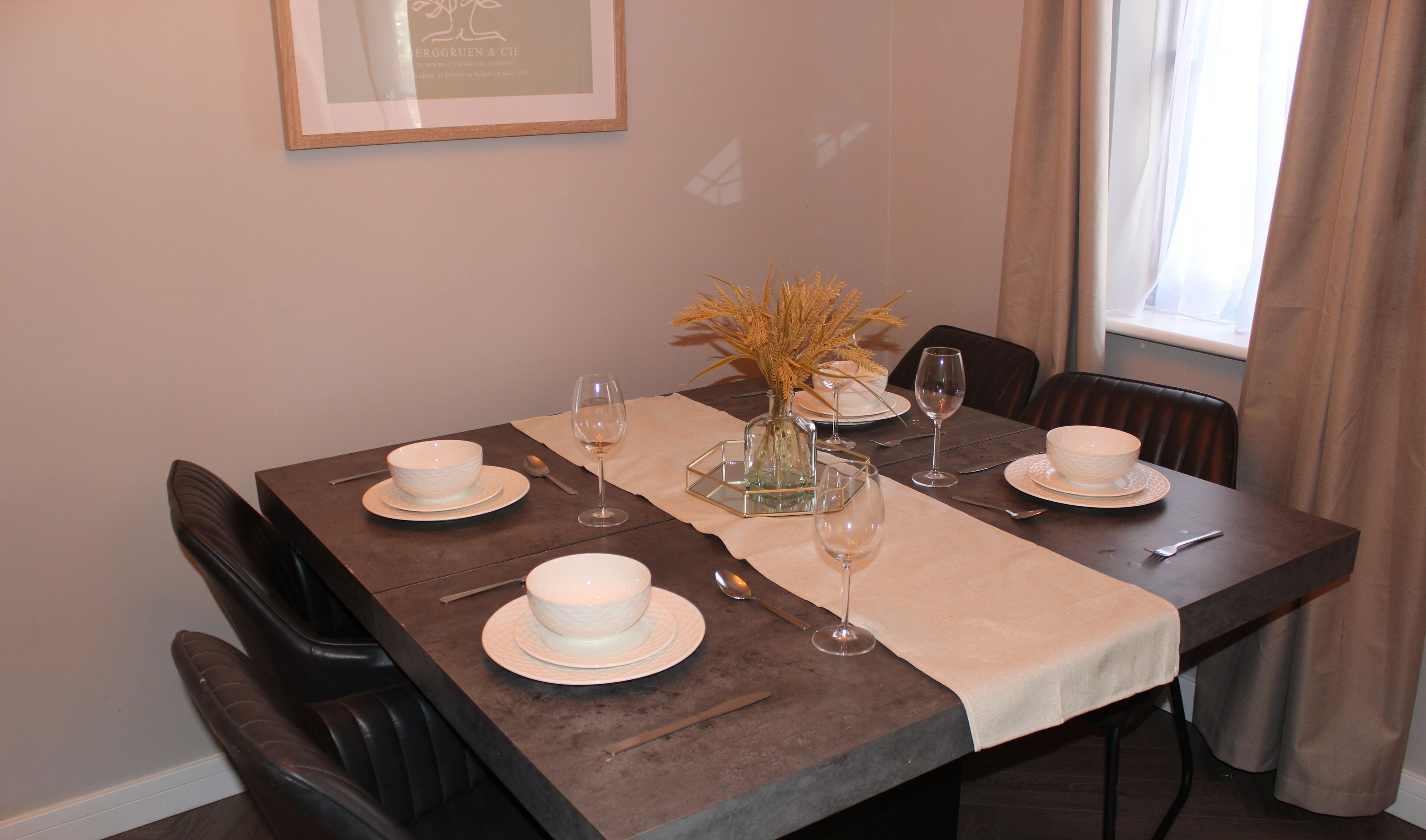 Une table à manger dressée avec de l’assiette blanche, des bols, des verres à vin et un vase contenant des plantes séchées.