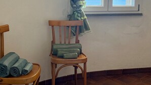 A room with a wooden chair, a small table with towels, a window with a view of the sea, and a curtain with a floral pattern. - Villa Gabriella "Sea View" (Upper Floor Apartment) (Santa Maria del Mare)