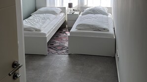 Une petite chambre à coucher moderne, avec deux lits simples, une table de chevet avec une lampe et une fenêtre agrémentée de rideaux légers.