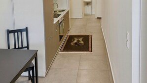 Un couloir étroit, avec une table et des chaises à manger sur la gauche, un coin cuisine avec des placards et un évier sur la droite, et une horloge au mur.