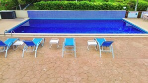 Une piscine à l’eau bleue, entourée de carreaux hexagonaux. On y trouve des chaises longues et une table recouverte d’un tapis bleu.