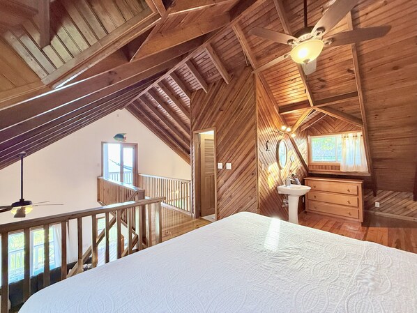 Une chambre mansardée avec un plafond haut, un plafond en bois, un ventilateur de plafond, un lit, une fenêtre avec des rideaux, une coiffeuse avec un lavabo et un escalier menant à l’étage supérieur.