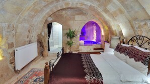 A stone-walled bedroom with a bed, a small sitting area, and a stone archway leading to a bathroom with a blue-lit sink. - Castle Inn Cave Cappadocia (Nevşehir)