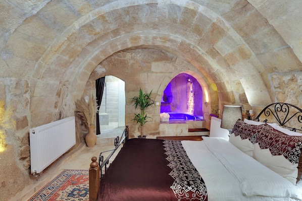 Un dormitorio de paredes de piedra con cama, una pequeña zona de estar y un arco de piedra que conduce a un baño con un lavamanos iluminado en azul.