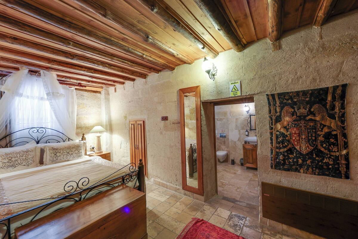 A bedroom with a wooden ceiling, a bed with a canopy, a bathroom visible through an open door, and a tapestry on the wall.