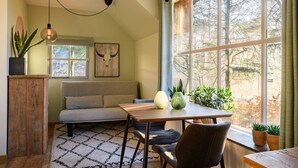 A cozy room with a sofa, a table, a hanging light, a window with a view of trees, and a plant on the windowsill. - Ultra-luxe Bungalow in Herperduin (Herpen)
