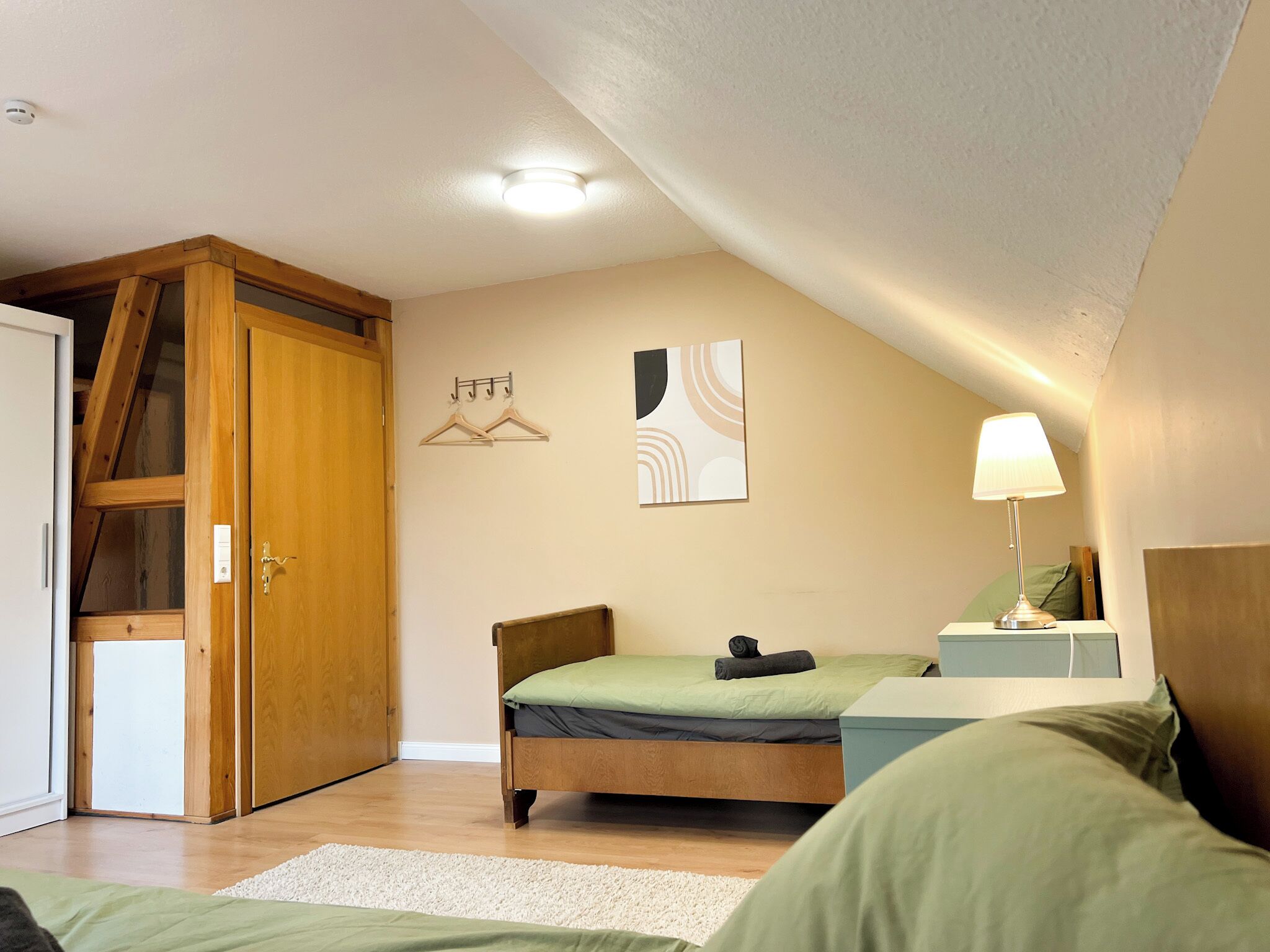 Une chambre avec une armoire en bois, un lit avec une literie verte, une table de chevet avec une lampe et un porte-manteau fixé au mur.