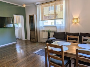 A modern living room with a wooden dining table, a sofa, and a large window with sheer curtains.