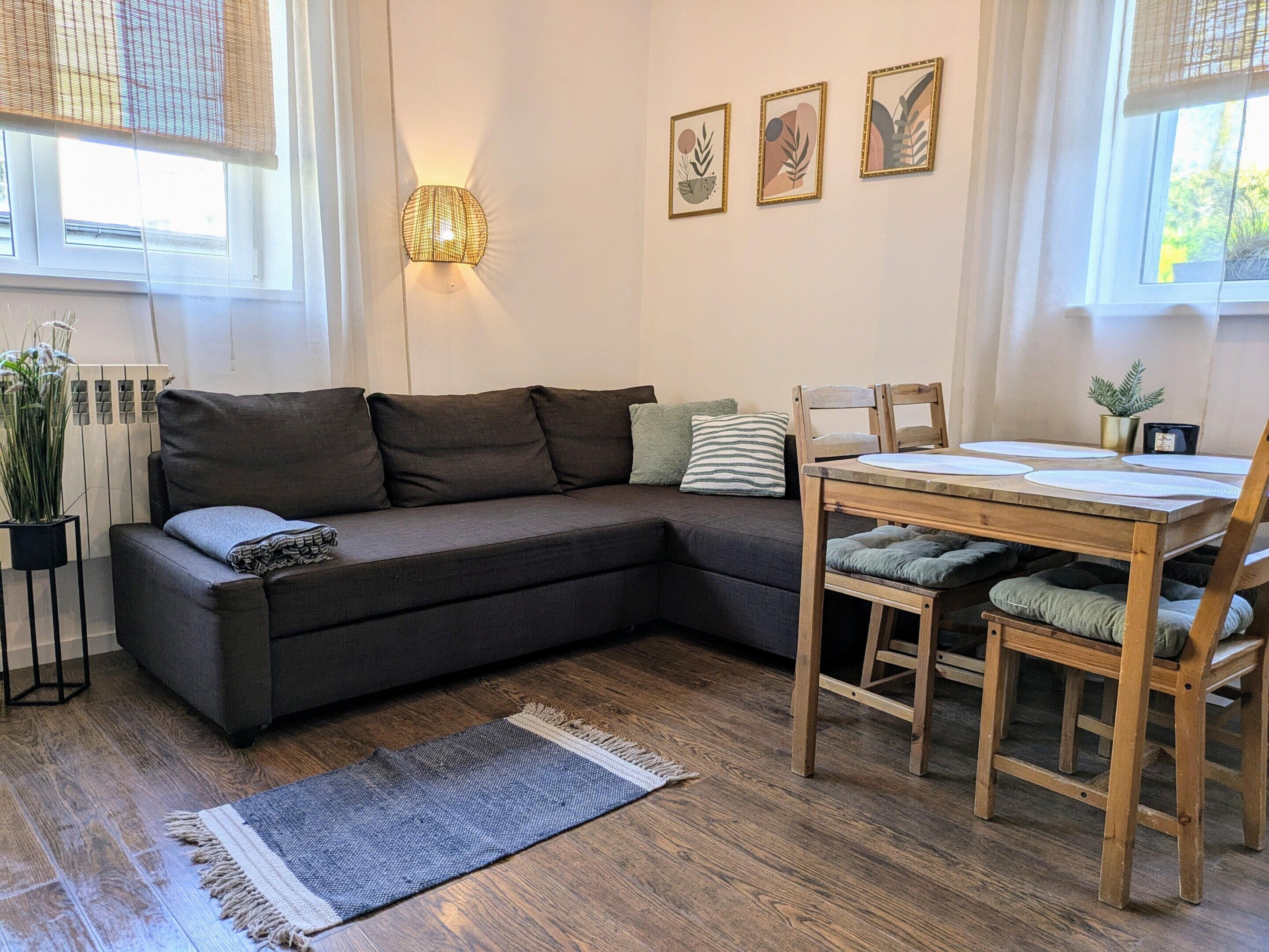 Ein modernes Wohnzimmer mit einem dunkelbraunen Ecksofa, einem Holzesstisch mit Stühlen, einer Stehlampe und drei gerahmten Kunstwerken an der Wand.