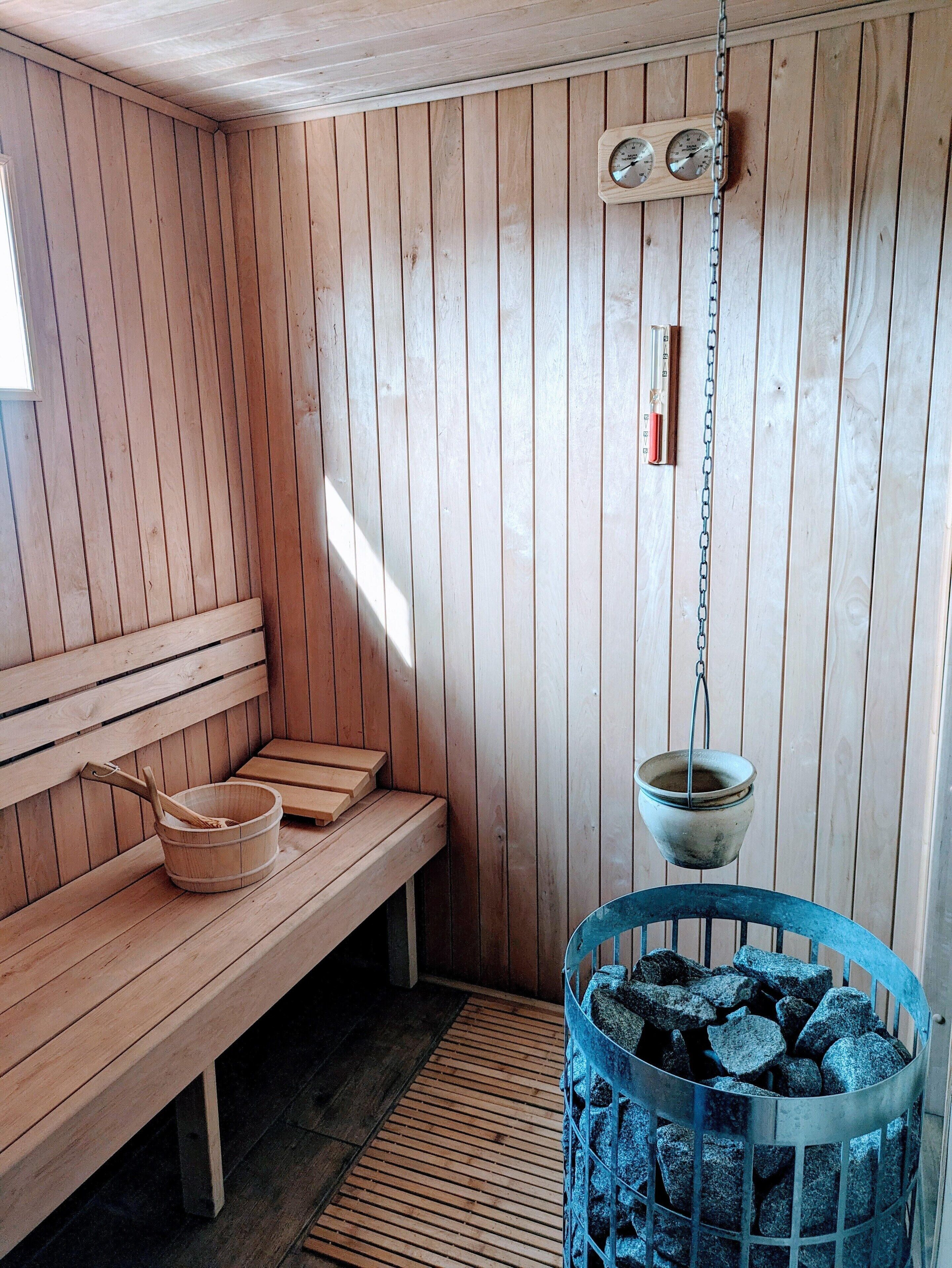 A sauna room with wooden benches, a bucket, and a metal basket of stones.