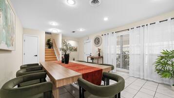 A dining area with a wooden table, green chairs, and a red runner. There are ceiling lights, a clock, and a window with curtains.