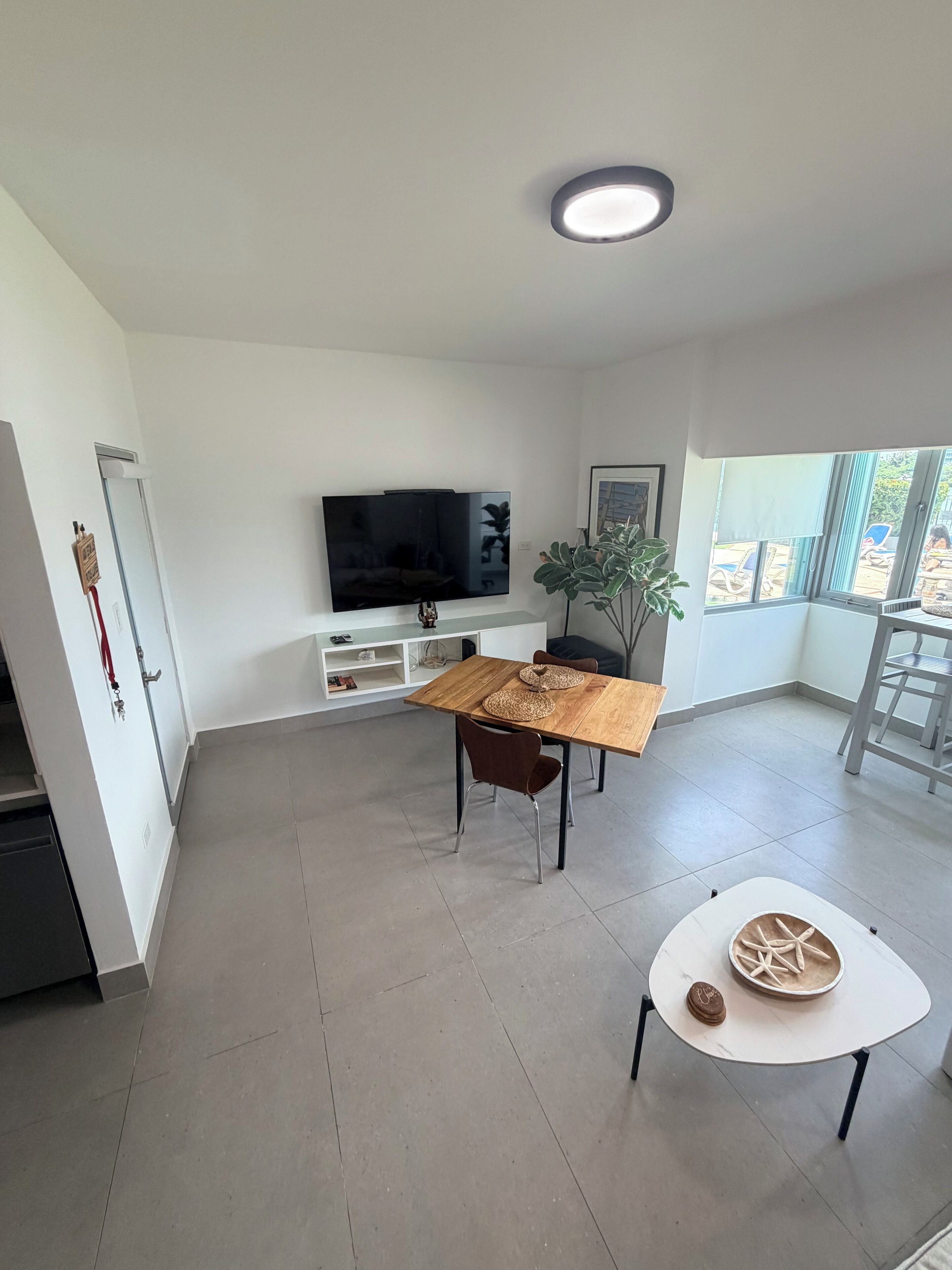 A modern living room with a flat-screen TV, a dining table with chairs, a small white table with decorative items, and a large window with a view of the outdoors.
