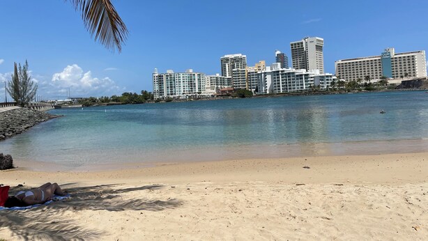 Ein Strand mit einer Stadtansicht im Hintergrund, darunter ein Hotel namens "Calle Del Aire".