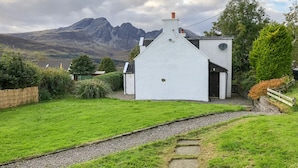 A white house with a steep roof, surrounded by a grassy area and a stone path leading to the entrance. - HAWTHORN DENE, pet friendly, cosy croft cottage with mountain views in Torrin (Torrin)