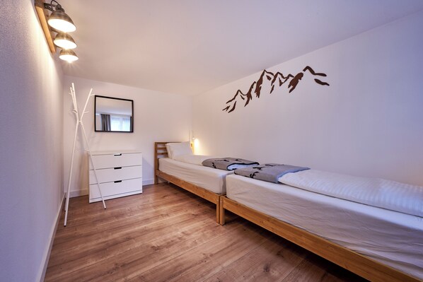 A small, neatly arranged bedroom with a wooden floor, a bed with a white mattress and a blanket, a bedside table with a lamp, a mirror on the wall, and a wall decoration of mountains.