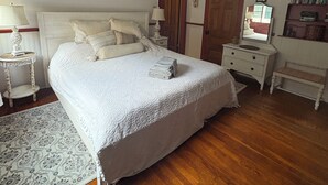 Een slaapkamer met een groot bed, een plafondventilator, een nachtkastje, een dressoir, een spiegel en een boekenplank.
