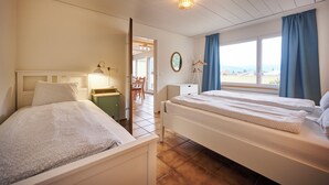 A bedroom with two beds, a desk, and a chair. There is a window with curtains and a view of the outside.