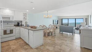 A modern kitchen with white cabinetry, a central island, and a dining area with wicker chairs. - Gulf-Front Condo - Heated Pool - Sunset Views - Direct Beach Access - Sleeps 5 (Indian Shores)