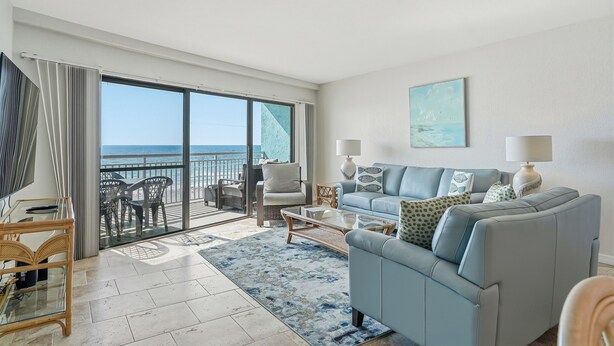 A spacious living room with a large sliding glass door leading to a balcony, a light blue sofa, a coffee table, and a television. - Gulf-Front Condo - Heated Pool - Sunset Views - Direct Beach Access - Sleeps 5 (Indian Shores)