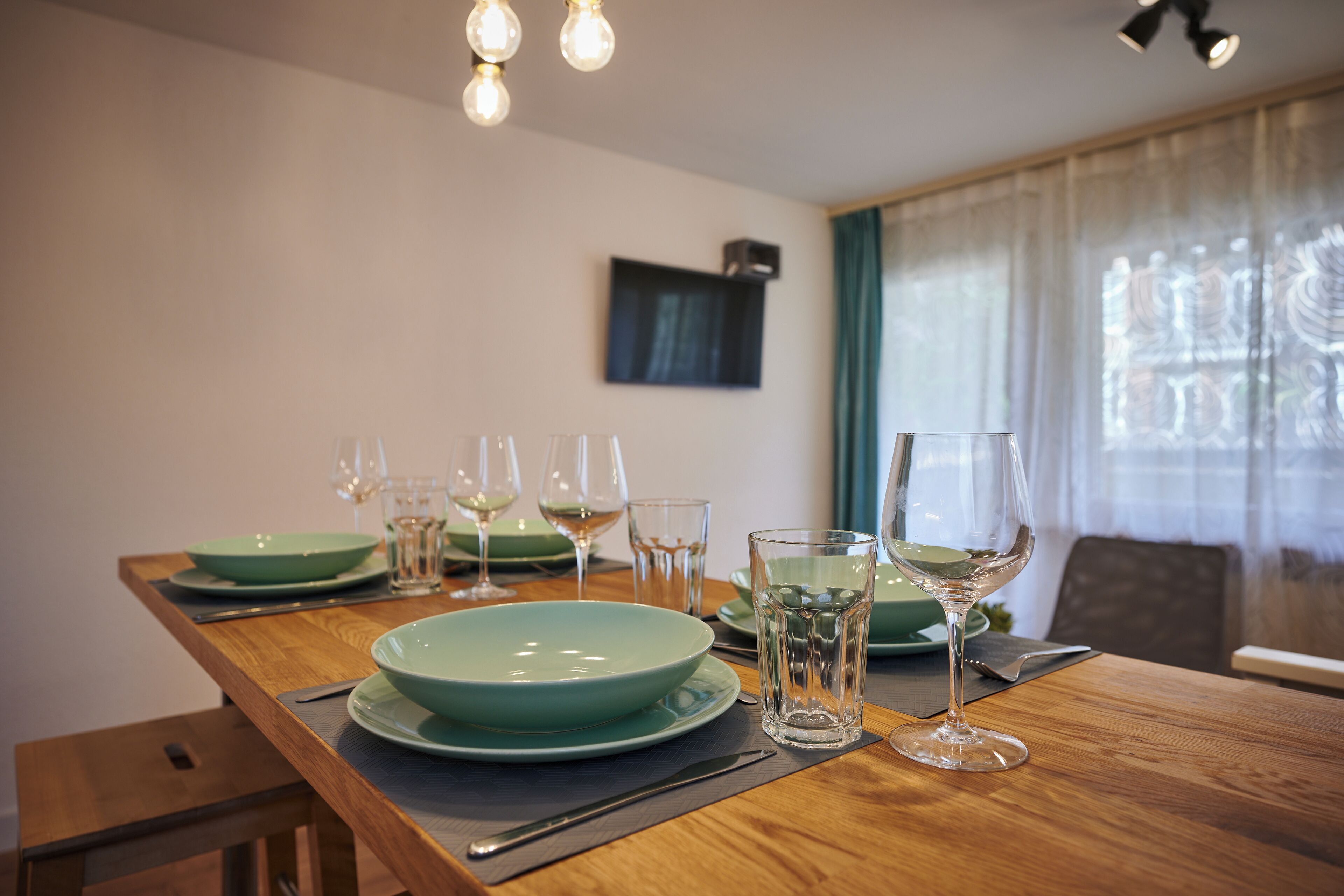 A dining table set with green plates, glasses, and cutlery, with a wooden table and a TV mounted on the wall.