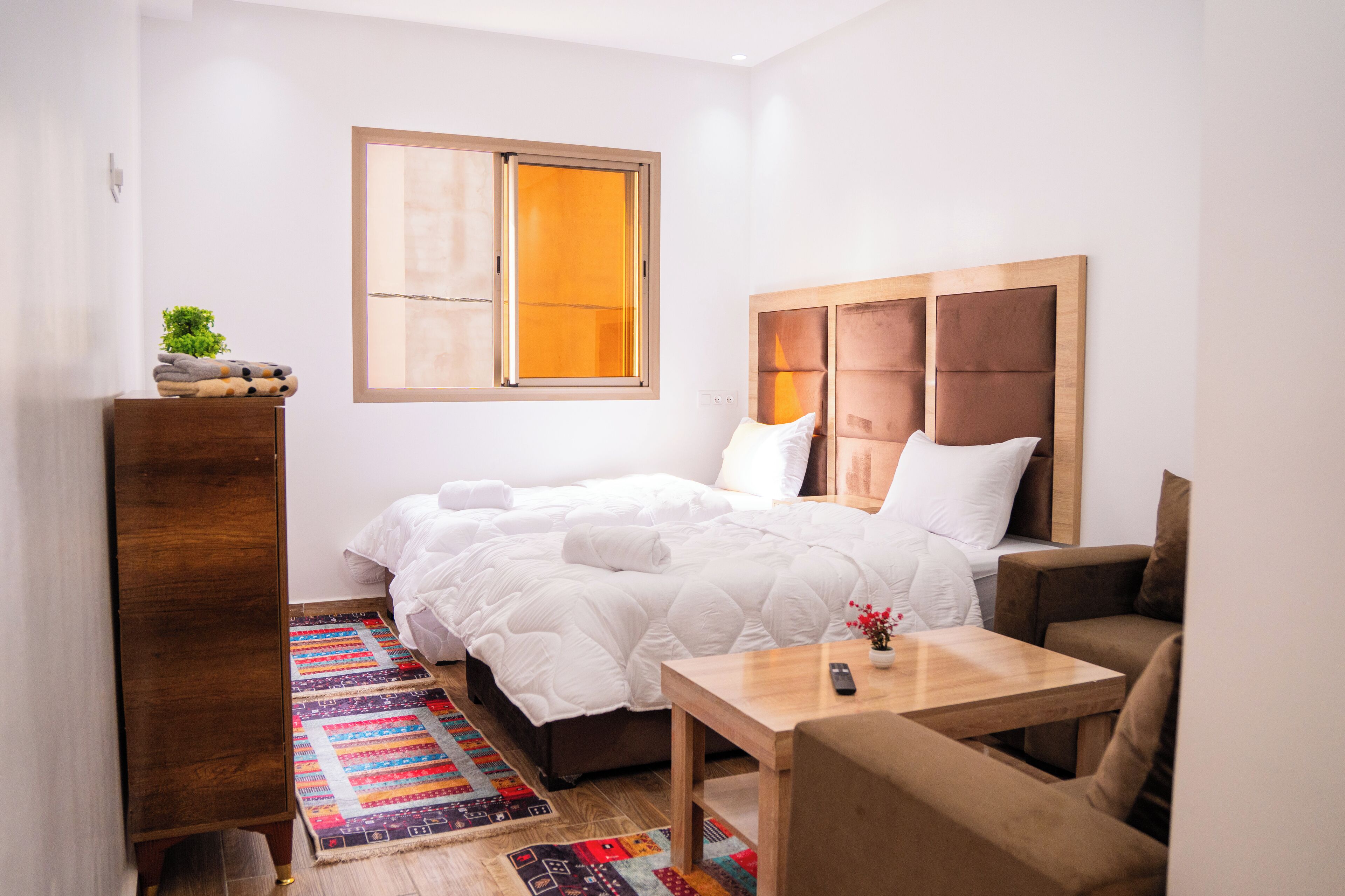 A bedroom with a bed, a wooden headboard, a small table, a couch, a window, and a colorful rug.