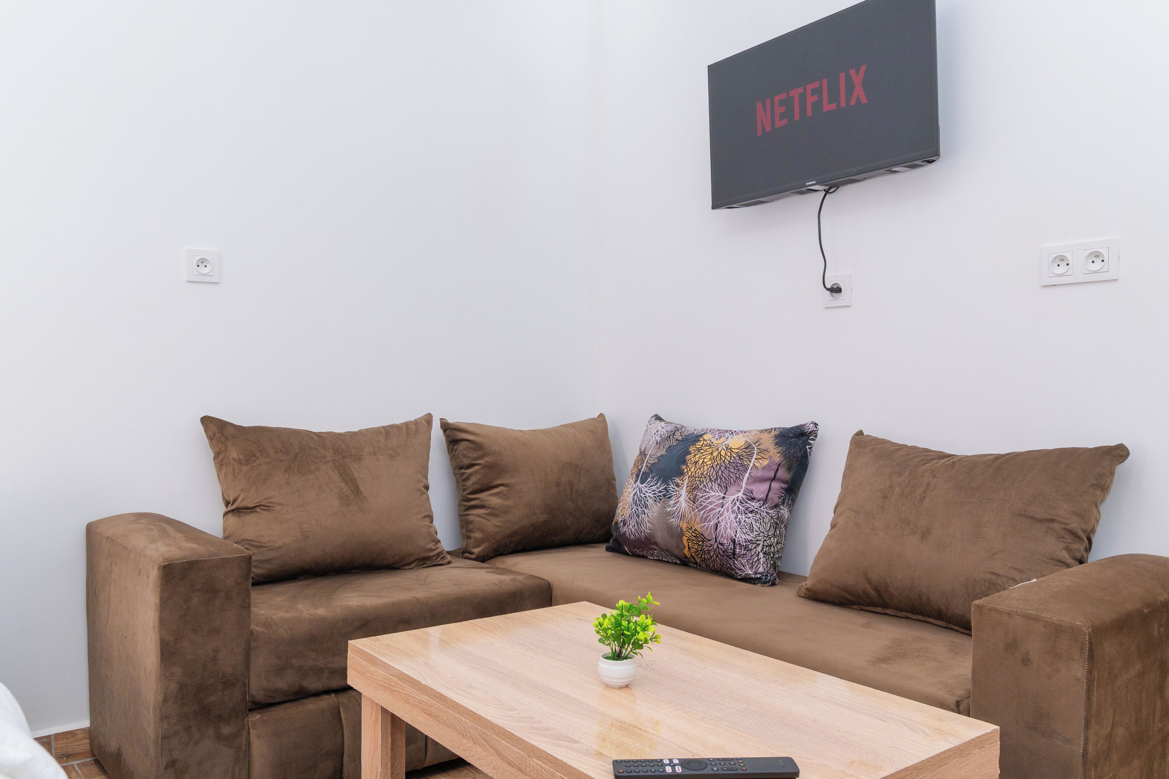 Ein modernes Wohnzimmer mit einem braunen Sofa, einem Holztisch und einem an der Wand befestigten Fernseher, der das Netflix-Logo zeigt.