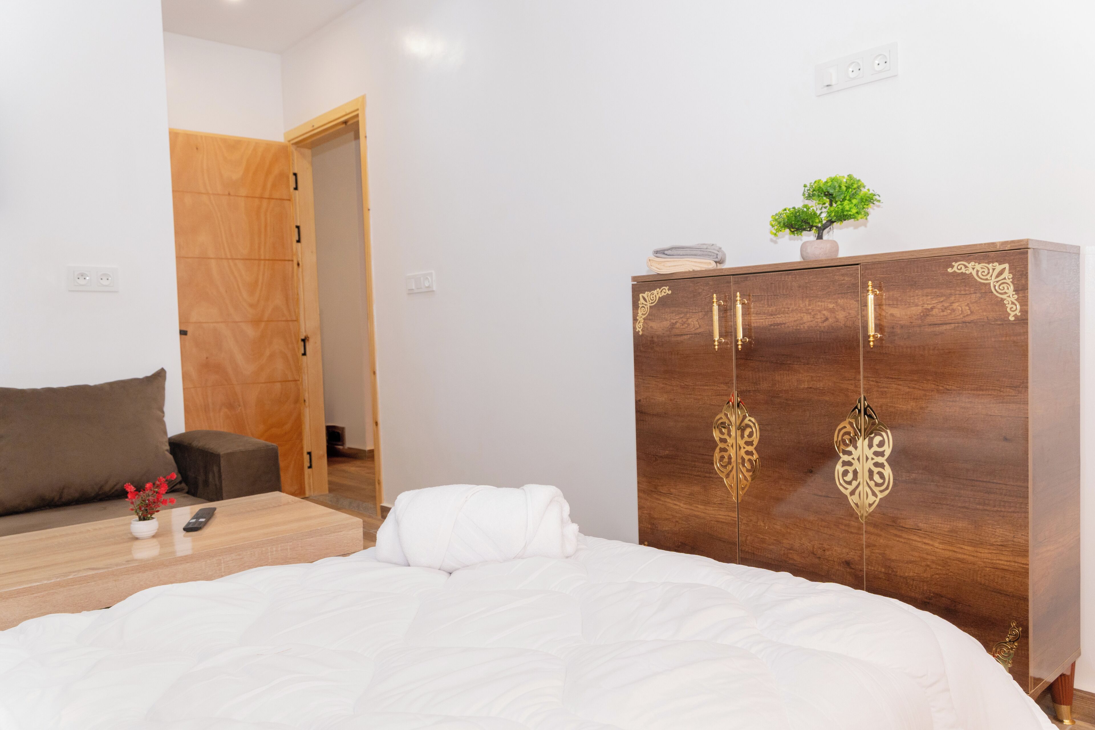 A bedroom with a wooden wardrobe, a bed with white bedding, a small table with a remote control, and a potted plant on the wardrobe.