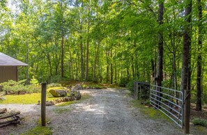 Ein Kiesweg schlängelt sich durch ein bewaldetes Gebiet, rechts befindet sich ein Metalltor.