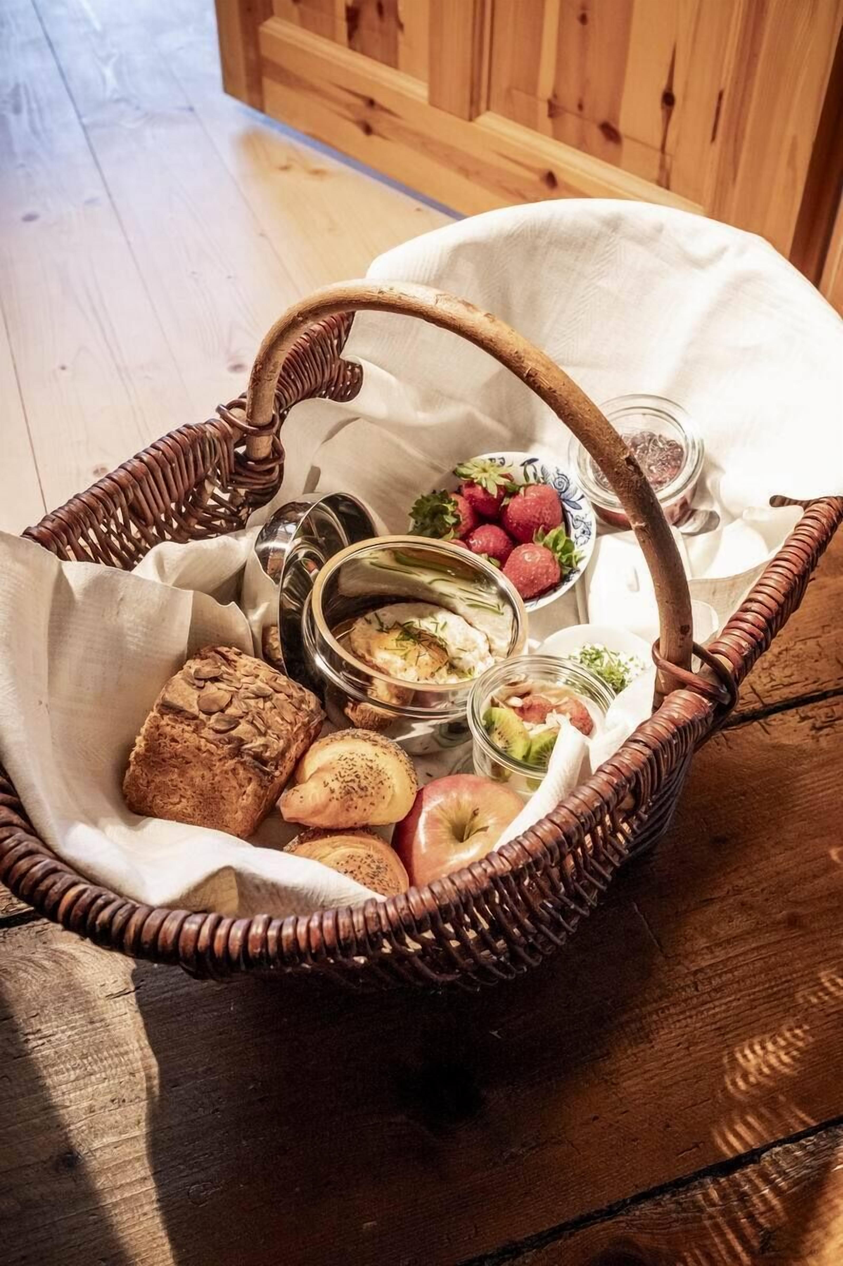En korg av flätat material, fylld med matvaror, inklusive bröd, frukt och en skål med jordgubbar.
