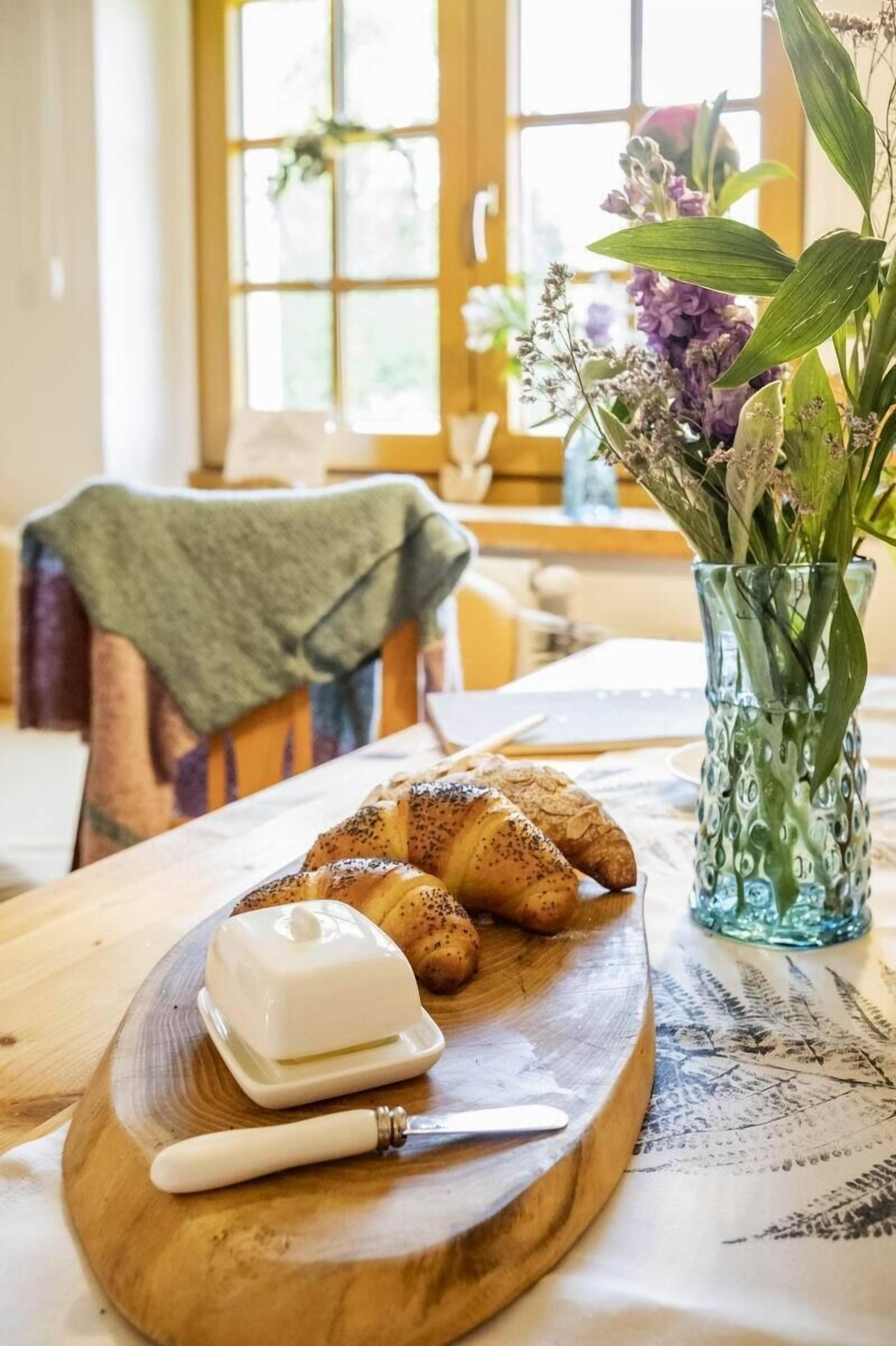 A wooden board with croissants and a butter knife on a table, a vase with flowers, and a window with a view of the outside.