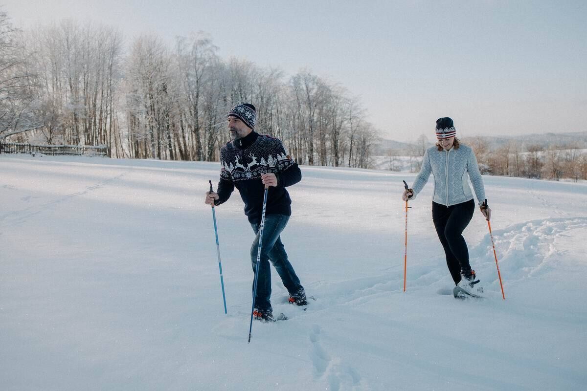Two people cross-country skiing in a snowy landscape with bare trees in the background.