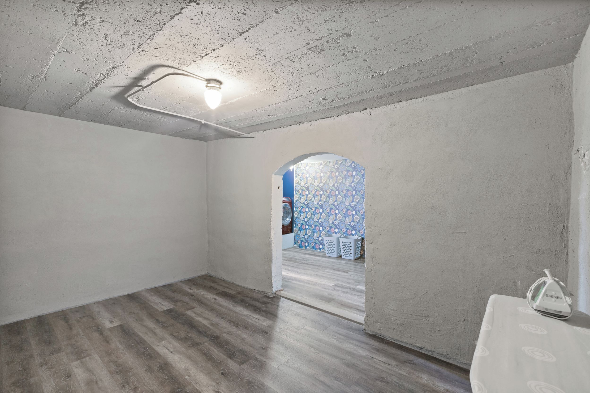 Une pièce dotée d’un plancher en bois, d’un luminaire au plafond et d’une porte-fenêtre donnant sur une autre pièce dont les murs sont recouverts d’un papier peint à motifs.