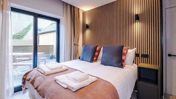 A modern hotel room with a large bed, a bedside table, and a view of the outdoors through a sliding glass door. - Modern 2-Bedroom Apartment with Parking (Conwy Principal Area)