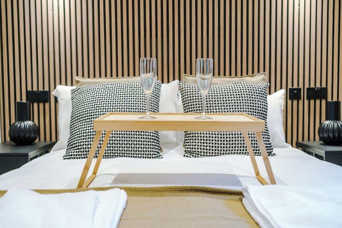 Ein Hotelzimmer mit einem Bett, ausgestattet mit schwarz-weiß gemusterten Kissen, einem Nachttisch aus Holz mit zwei Gläsern Wasser und einem kleinen Holztablett auf dem Bett.