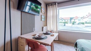 A modern room with a dining table set for two, a flat-screen TV mounted on the wall, and a large window with a view of houses.