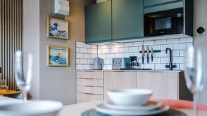 A modern kitchen with white tiles, a microwave, and a coffee machine. There are framed artworks on the wall and a dining table with plates and glasses.