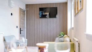 Una habitación de hotel moderna con zona de comedor, televisor montado en una pared con listones y una planta pequeña en el alféizar.