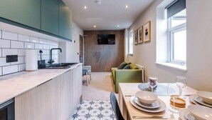 Una cocina moderna con una isla central, un comedor con una mesa preparada para cuatro personas y una sala de estar con un sofá y televisor.