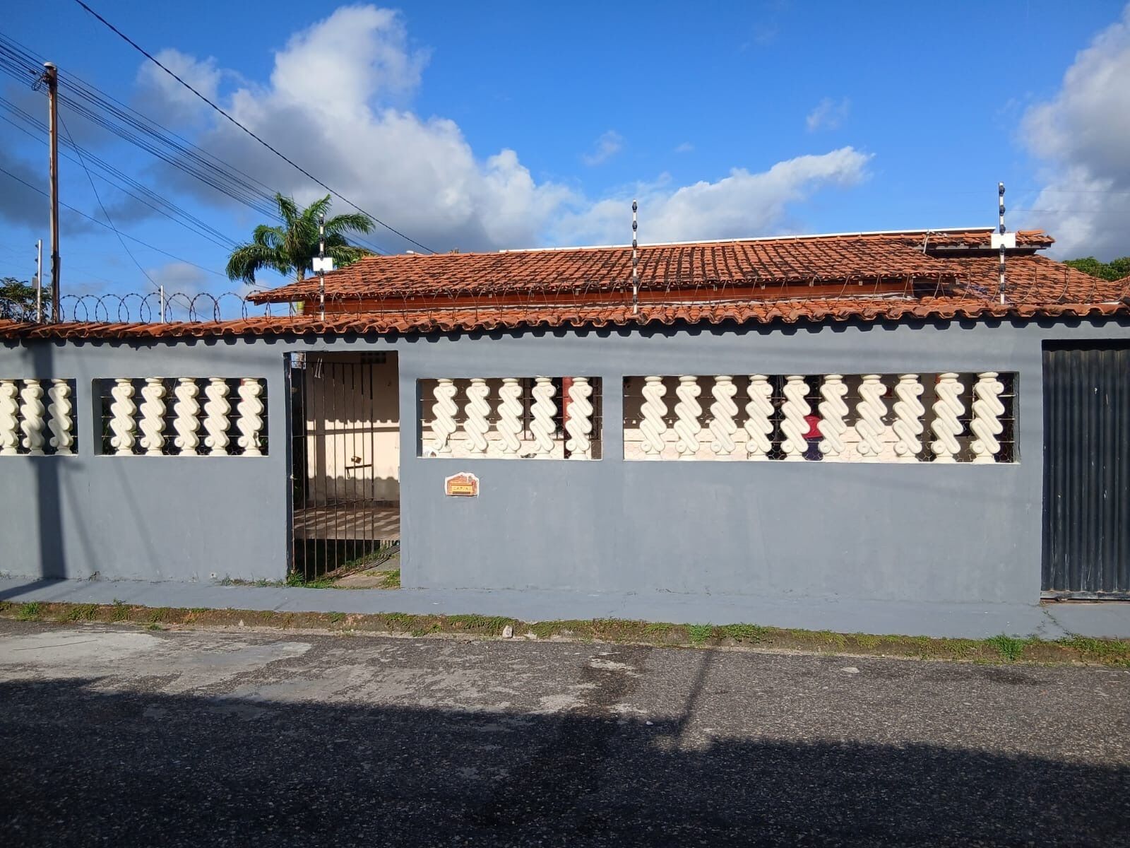 Un bâtiment gris avec un toit recouvert de tuiles, une entrée fermée par un portail et une petite enseigne sur le mur.